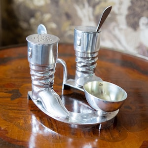 May include: A silver-colored salt and pepper set and a small bowl. The salt and pepper shakers are shaped like boots, with a small spoon in one. The set is on a decorative tray.