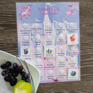 May include: A vibrant 'Annabel's Foodie BINGO' game card with a unicorn and fairy theme. The card features squares with phrases like 'Butterfly brilliance!' and 'Unicorn triumph!' alongside fruit illustrations. A bowl of blackberries, blueberries, and a lime is in the foreground.