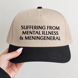 May include: A beige baseball cap with a black brim and a black button on top. The front of the cap features black embroidered text that reads: SUFFERING FROM MENTAL ILLNESS & MENINGENERAL. The cap has a structured crown and a flat, solid black visor.
