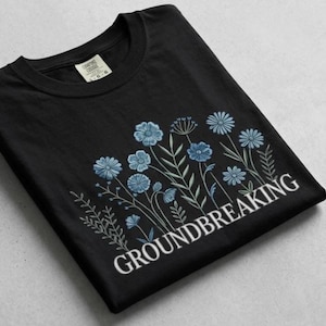 Puede incluir: Camiseta negra con un diseño floral en tonos azules y verdes. La palabra "GROUNDBREAKING" está impresa en blanco debajo de las flores. La camiseta está doblada y tiene una etiqueta en la esquina superior izquierda.
