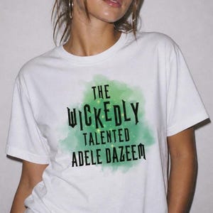 Może przedstawiać: Biały t-shirt z krótkim rękawem z zieloną grafiką w stylu akwareli na klatce piersiowej. Napis THE WICKEDLY TALENTED ADELE DAZEEM wykonano czcionką szeryfową w kolorze czarnym, z drobnymi żółtymi iskierkami wkomponowanymi w zielony wzór.