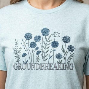 Peut inclure: Un t-shirt chiné bleu clair avec une broderie bleu foncé représentant diverses fleurs sauvages et tiges. Le mot "GROUNDBREAKING" est brodé dans une police serif bleu foncé juste en dessous du motif floral sur le devant du t-shirt.