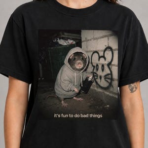 May include: A black t-shirt featuring a graphic of a rat wearing a gray hoodie and holding a spray paint can. The rat is standing next to a brick wall with a mouse-shaped graffiti design. Below the image, the text reads: it's fun to do bad things.