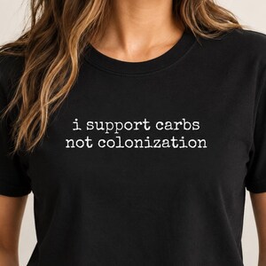 Può includere: Una t-shirt nera a maniche corte con testo bianco stampato sul petto che recita "i support carbs not colonization" in un carattere in stile macchina da scrivere. Il capo ha uno scollo rotondo e una vestibilità standard in tessuto nero.