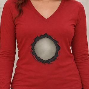 Pode incluir: Uma camisola vermelha de manga comprida com decote em V, apresentando um design gráfico central que imita um efeito de tecido rasgado, revelando um núcleo circular claro e texturizado rodeado por bordas escuras, irregulares e desgastadas.