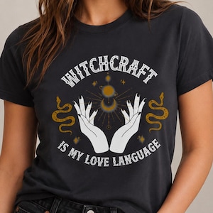 Puede incluir: Una camiseta gris carbón de manga corta con un estampado gráfico de dos manos extendidas hacia un símbolo celestial de sol y luna. El texto Witchcraft is my love language está flanqueado por dos serpientes doradas.