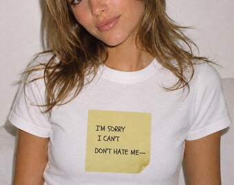 May include: White t-shirt with a yellow sticky note graphic. The note reads "I'M SORRY I CAN'T DON'T HATE ME-" in black text. The shirt has short sleeves and a crew neck.