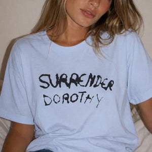 以下が含まれることがあります： 胸元に黒の手書き風フォントで「SURRENDER DOROTHY」というテキストがプリントされた、ライトブルーの半袖Tシャツ。この衣類は、クラシックなクルーネックとリラックスしたフィット感が特徴で、無地の生地で作られています。