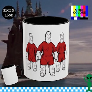Könnte beinhalten: Weißer Keramikbecher mit schwarzem Innenraum und Griff. Der Becher zeigt ein skurriles Design von drei fingerförmigen Figuren, die rote Hemden und Shorts tragen. Der Text "11oz & 15oz" befindet sich oben links. Die Worte "MORE COLORS" stehen oben rechts.