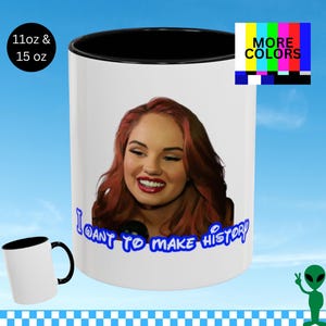 Könnte beinhalten: Weißer Keramikbecher mit schwarzem Innenraum und Griff. Der Becher zeigt ein Porträt einer Frau mit roten Haaren und den blauen Text "I WANT TO MAKE HISTORY". Ein kleinerer Becher ist unten links sichtbar.