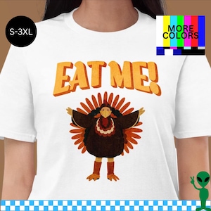 May include: White t-shirt with the text "EAT ME!" in orange letters above a cartoon turkey with outstretched wings. The turkey is brown with orange and red accents. The shirt is available in sizes S-3XL.