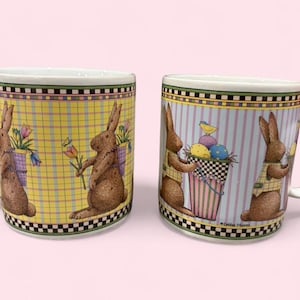 May include: Two ceramic Easter mugs with white handles. One mug has a yellow plaid pattern with brown bunnies carrying flowers. The other mug has a striped pattern with bunnies carrying Easter eggs. Both mugs have a decorative border.