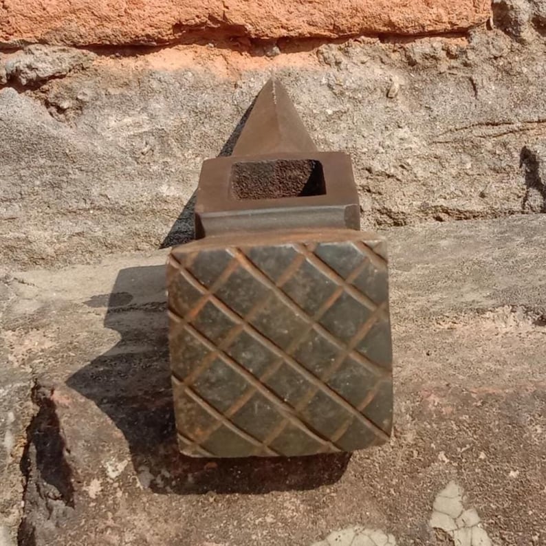 May include: A weathered, dark brown metal object with a geometric design. The object has a square base with a diamond pattern, a rectangular midsection, and a pyramidal top. The object appears to be made of iron or a similar metal.