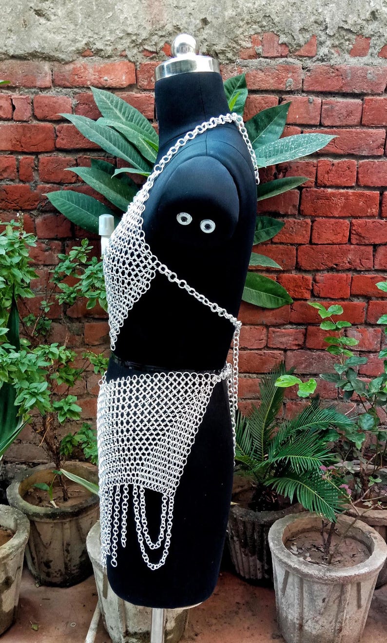 May include: A silver chainmail top and skirt set displayed on a black mannequin. The chainmail is made of small, interlocking metal rings. The top is a halter style, and the skirt has a fringed hem. The background features a brick wall and greenery.