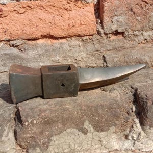 May include: A vintage, weathered metal anvil with a rectangular body and a pointed horn. The anvil is a dark, muted gray color, showing signs of age and use. It rests on a rough, textured surface.