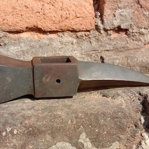 May include: A weathered, dark metal axe head with a rectangular eye and a long, pointed blade. The axe head is resting on a rough, textured stone surface, with a brick wall in the background.