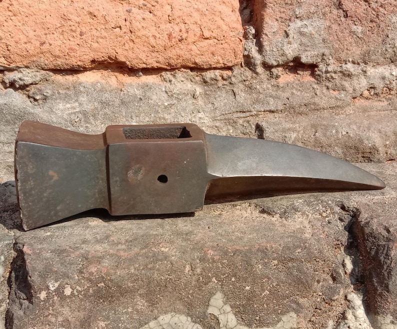 May include: A weathered, dark metal axe head with a rectangular eye and a long, pointed blade. The axe head is resting on a rough, textured stone surface, with a brick wall in the background.