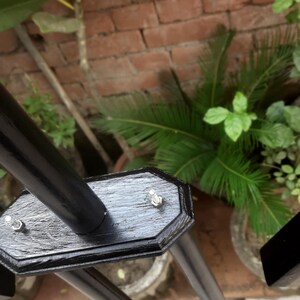 May include: Black metal frame with a rectangular base and a series of vertical supports. The frame is attached with silver bolts. The background features a brick wall and green plants.