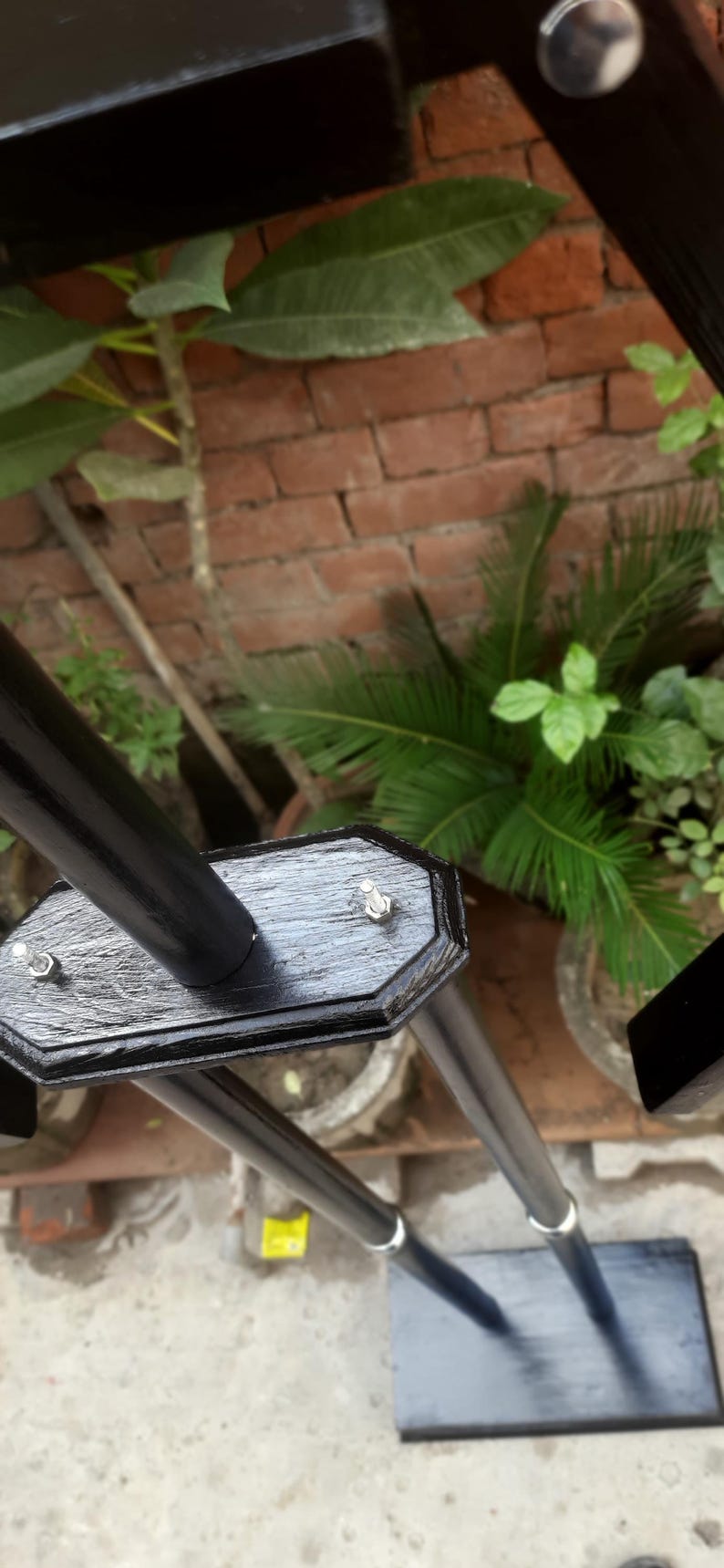 May include: Black metal frame with a rectangular base and a series of vertical supports. The frame is attached with silver bolts. The background features a brick wall and green plants.