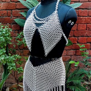 May include: A silver chainmail bikini set displayed on a black mannequin. The set includes a halter top and a skirt with a fringed hem. The mannequin is set against a backdrop of a brick wall and greenery.