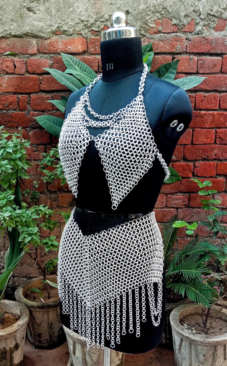 May include: A silver chainmail bikini set displayed on a black mannequin. The set includes a halter top and a skirt with a fringed hem. The mannequin is set against a backdrop of a brick wall and greenery.