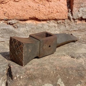 May include: A vintage, weathered metal tool with a textured, diamond-patterned striking face. The tool has a square socket and a pointed end, likely used for shaping or working with materials. The tool is a dark, rusty brown color.