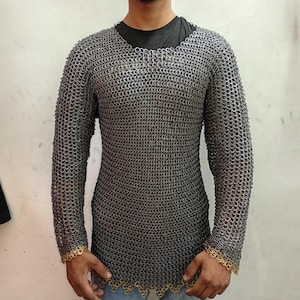 Puede incluir: Una camisa de cota de malla de manga larga hecha de pequeños anillos metálicos entrelazados. La camisa tiene un cuello redondo y llega hasta la mitad del muslo. Los puños y el dobladillo están adornados con un borde decorativo de color dorado. Se usa sobre una camiseta negra.
