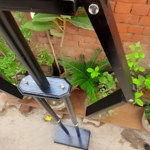 May include: A black wooden adjustable drafting table with a rectangular base and a hexagonal top. The table has a dark finish and is set against a brick wall with green plants.
