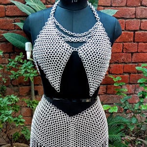 May include: A silver chainmail bikini set displayed on a black mannequin. The set includes a halter top with a chain necklace and a skirt with a fringed hem. The mannequin is set against a brick wall and greenery.