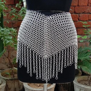 May include: A silver chainmail skirt displayed on a black mannequin. The skirt is made of small, interconnected metal rings, with a fringed bottom edge. The background features a brick wall and potted plants.