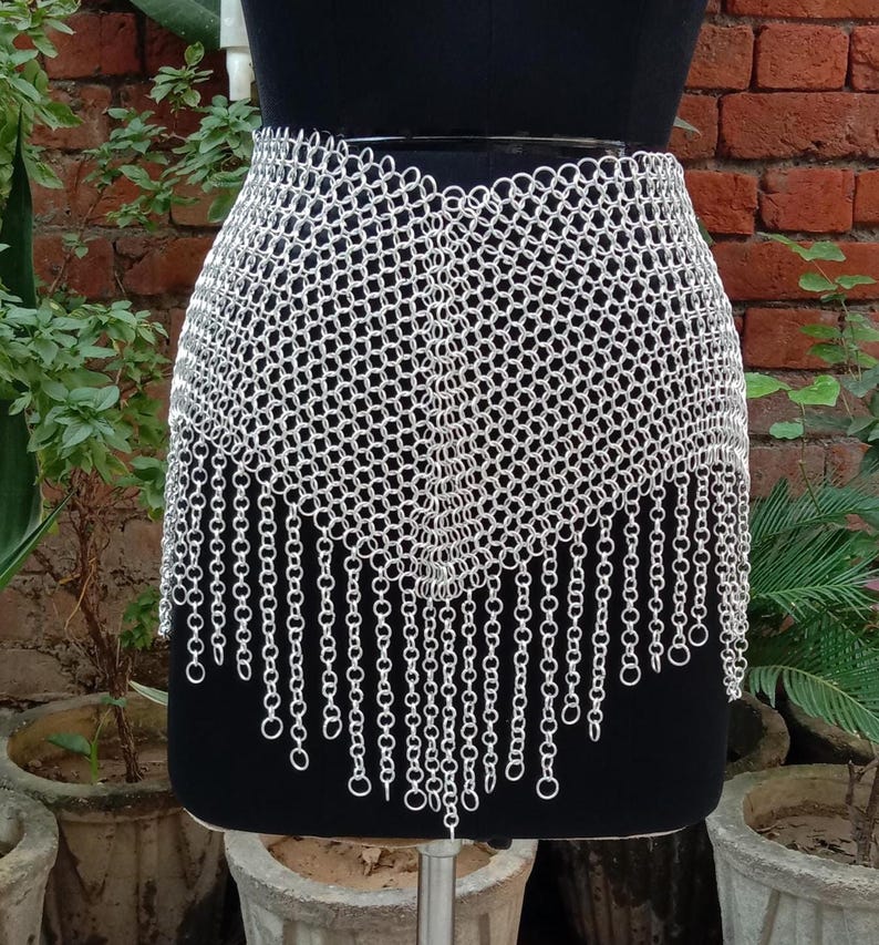 May include: A silver chainmail skirt displayed on a black mannequin. The skirt is made of small, interconnected metal rings, with a fringed bottom edge. The background features a brick wall and potted plants.