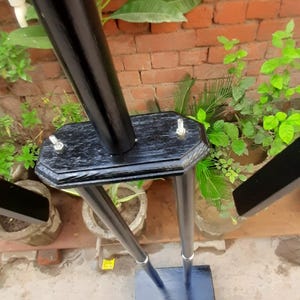 May include: A black, wooden plant stand with a hexagonal top and three legs. The stand has a rectangular base and is secured with silver bolts. The background features a brick wall and greenery.