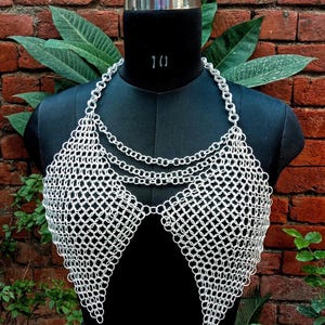 May include: A silver chainmail top displayed on a black mannequin. The top features a halter-style neckline with multiple chain strands and a diamond-patterned bodice. The background includes a brick wall and green foliage.