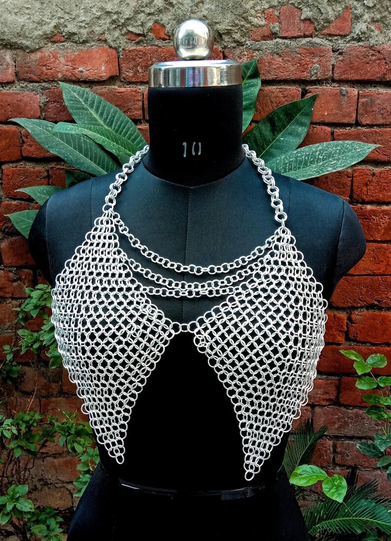 May include: A silver chainmail top displayed on a black mannequin. The top features a halter-style neckline with multiple chain strands and a diamond-patterned bodice. The background includes a brick wall and green foliage.