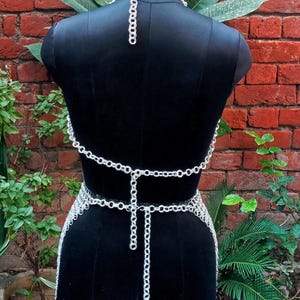 May include: A silver-colored chainmail top and waist belt displayed on a black mannequin. The chainmail is made of small, interlocking rings. The mannequin is set against a red brick wall with green plants in the background.