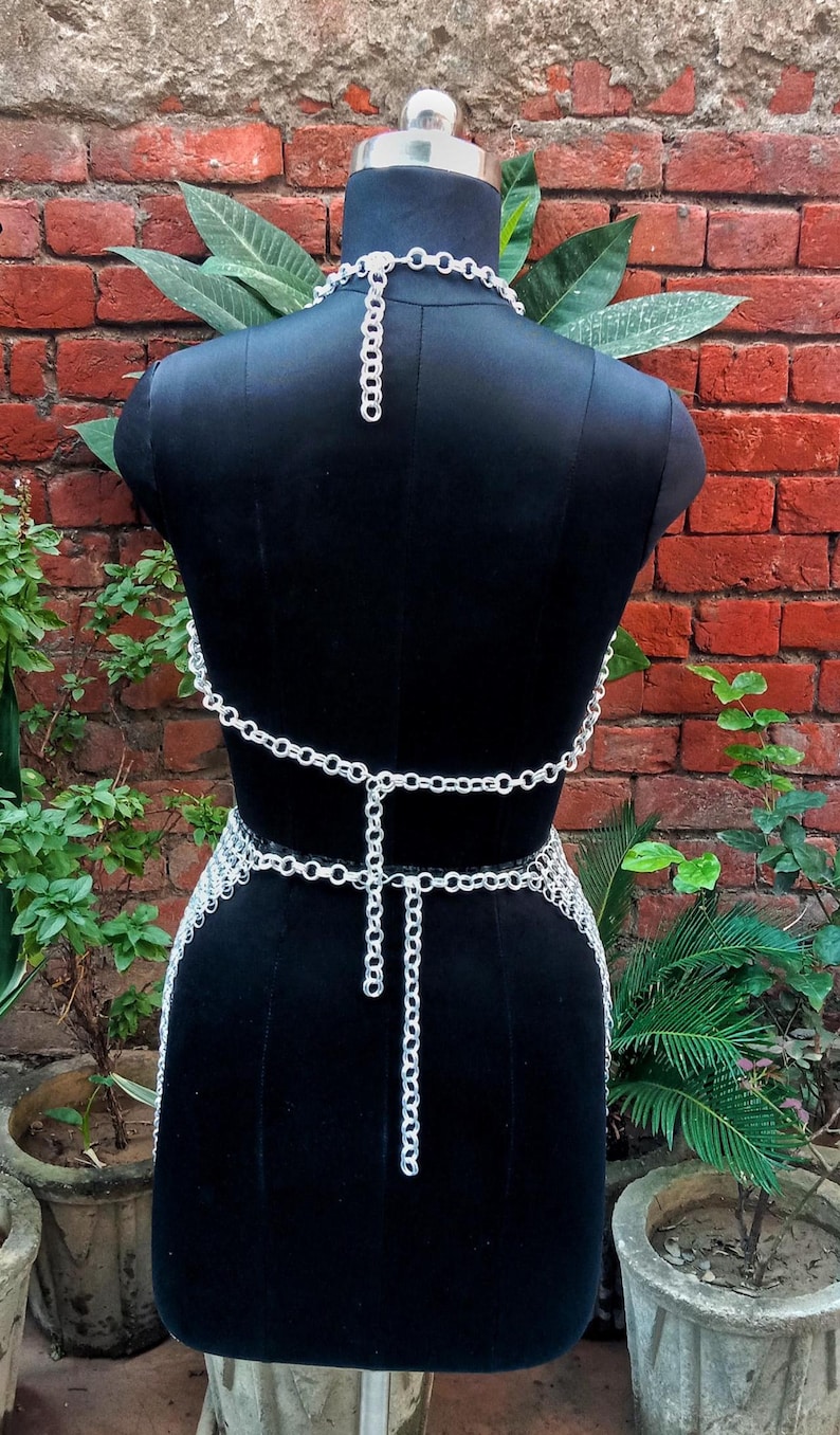 May include: A silver-colored chainmail top and waist belt displayed on a black mannequin. The chainmail is made of small, interlocking rings. The mannequin is set against a red brick wall with green plants in the background.