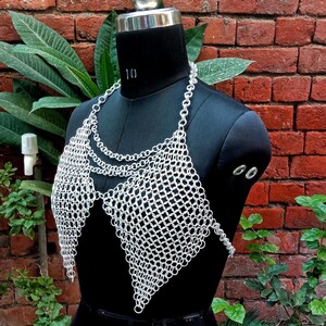 May include: A silver chainmail top displayed on a black mannequin. The top features a halter neck and a diamond-shaped design over the chest. The chainmail is made of small, interlocking metal rings. The background is a brick wall and green foliage.