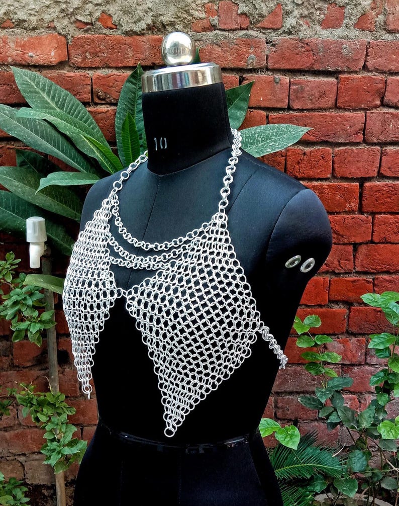 May include: A silver chainmail top displayed on a black mannequin. The top features a halter neck and a diamond-shaped design over the chest. The chainmail is made of small, interlocking metal rings. The background is a brick wall and green foliage.