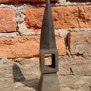 May include: A dark gray metal tool with a pointed top and a square opening in the middle. The tool has a rectangular base and appears to be made of iron or steel. The background is a weathered brick wall.
