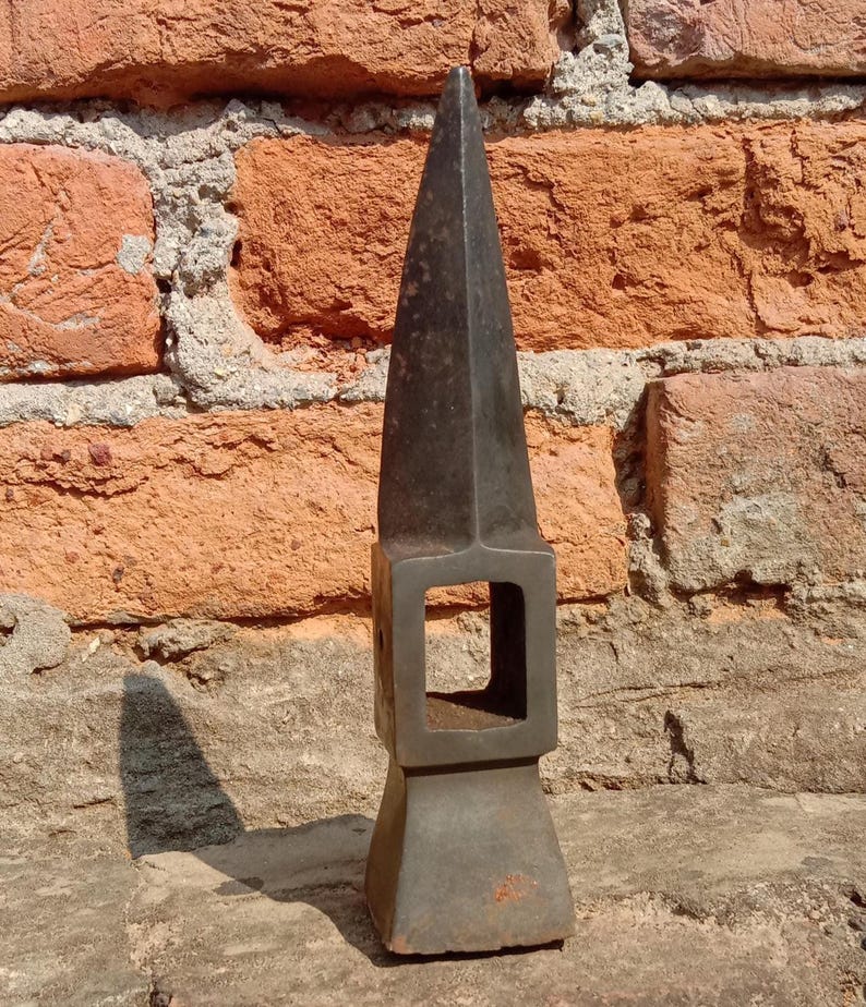 May include: A dark gray metal tool with a pointed top and a square opening in the middle. The tool has a rectangular base and appears to be made of iron or steel. The background is a weathered brick wall.