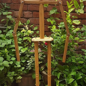 May include: A wooden artist's easel with adjustable arms and legs. The easel is made of light-colored wood and has a rectangular base. The background features lush green plants and a brick wall.