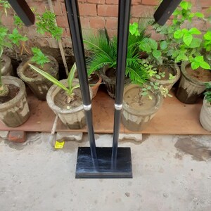 May include: A black metal stand with a rectangular base and two vertical supports. The stand has a top section with four angled arms. The background includes potted plants and a brick wall.