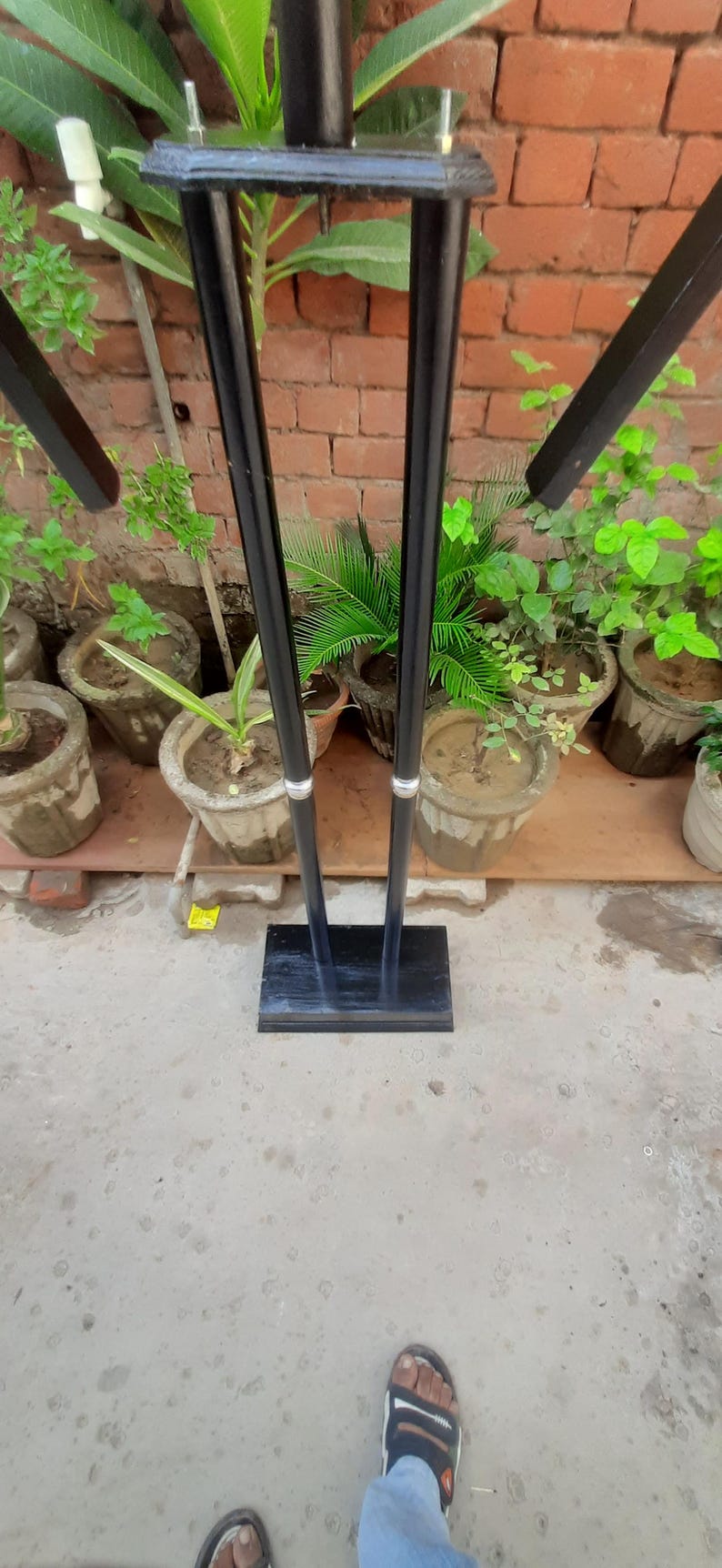 May include: A black metal stand with a rectangular base and two vertical supports. The stand has a top section with four angled arms. The background includes potted plants and a brick wall.