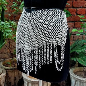 May include: A silver chainmail skirt with a draped design and dangling chain fringe. The skirt is displayed on a black mannequin, showcasing its intricate metalwork and texture. The skirt is a unique piece of apparel.