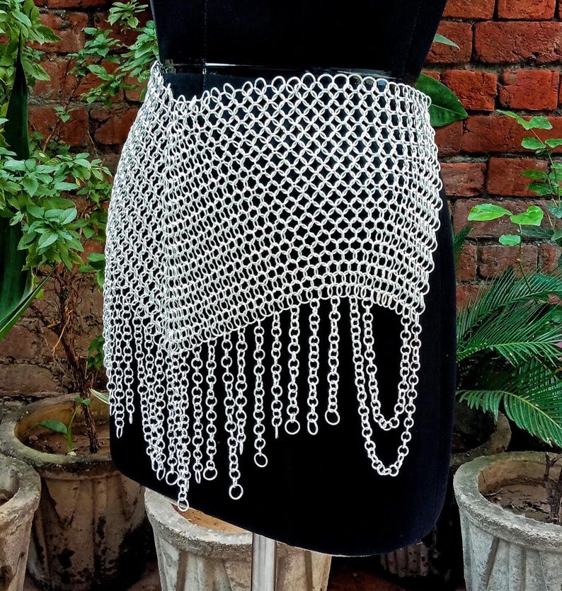 May include: A silver chainmail skirt with a draped design and dangling chain fringe. The skirt is displayed on a black mannequin, showcasing its intricate metalwork and texture. The skirt is a unique piece of apparel.