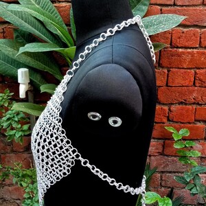 May include: A silver chainmail top displayed on a black mannequin. The top is made of small, interconnected metal rings, forming a mesh pattern. The chainmail extends over the shoulders and around the waist, with a chain closure. The background features a brick wall and green foliage.
