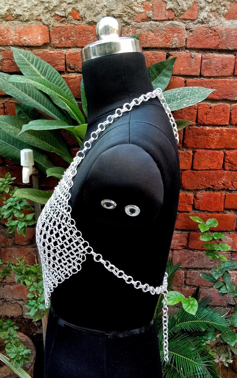 May include: A silver chainmail top displayed on a black mannequin. The top is made of small, interconnected metal rings, forming a mesh pattern. The chainmail extends over the shoulders and around the waist, with a chain closure. The background features a brick wall and green foliage.
