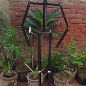 May include: A black metal mannequin stand with articulated arms and legs. The stand has a rectangular base and a round head. It is positioned in front of a brick wall and potted plants.
