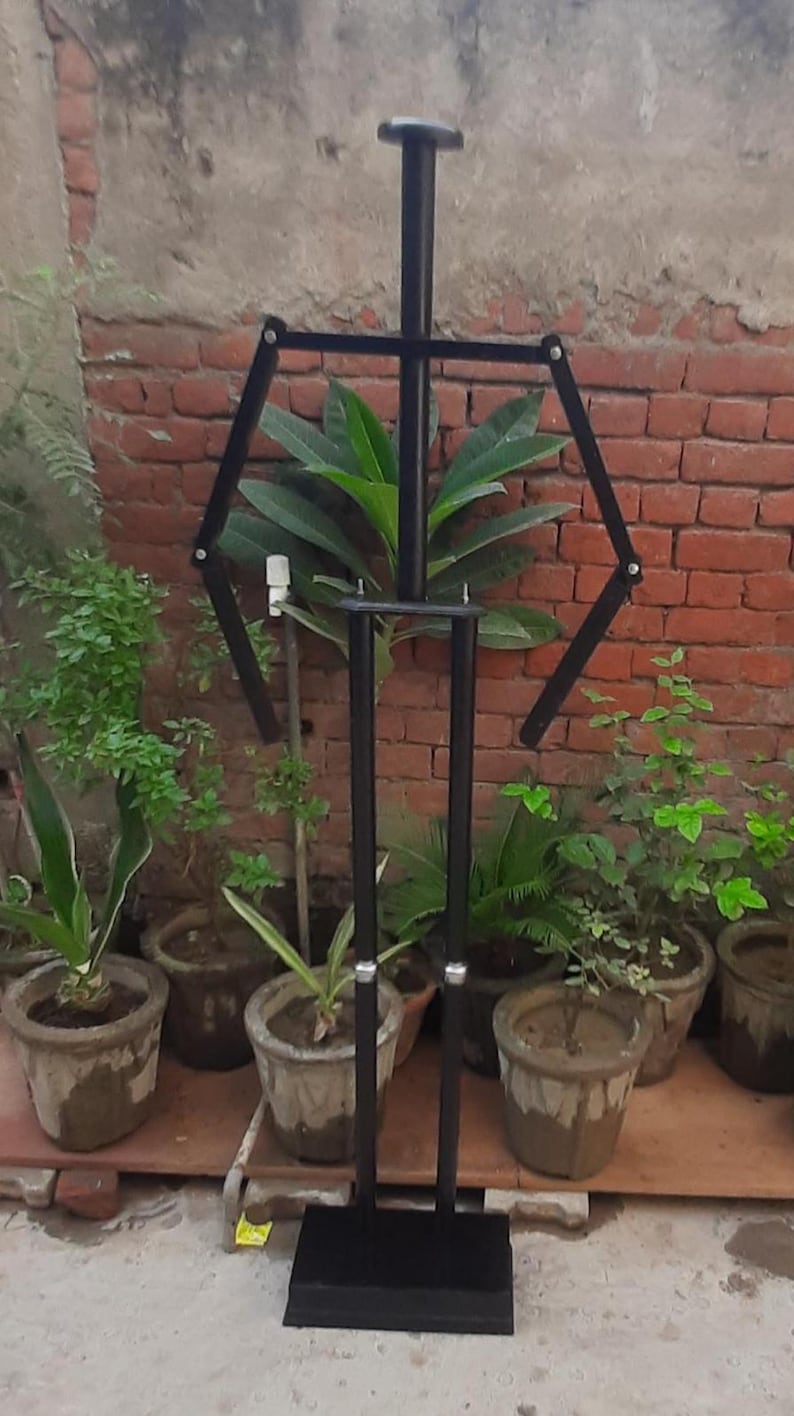May include: A black metal mannequin stand with articulated arms and legs. The stand has a rectangular base and a round head. It is positioned in front of a brick wall and potted plants.