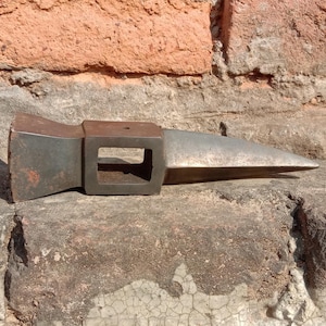May include: A weathered, metal tool head with a rectangular eye and a pointed, wedge-shaped blade. The tool has a dark, aged appearance, suggesting it's been used. The background features a rough, textured brick wall.